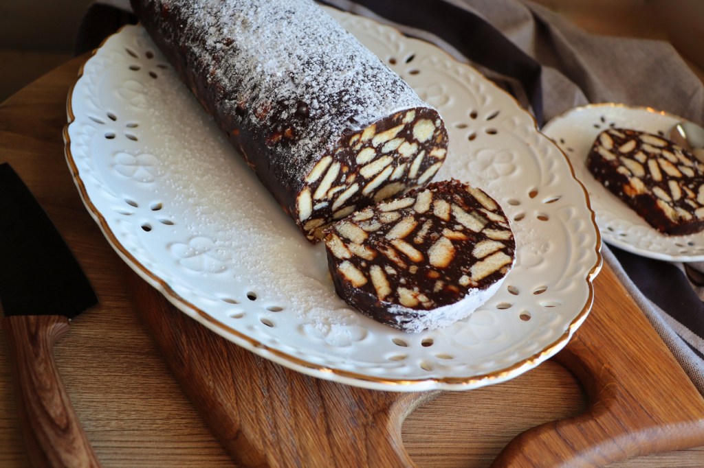 A close-up view of a chocolate salami with visible pieces of biscuits, dusted with powdered sugar, served on a white, ornate plate.