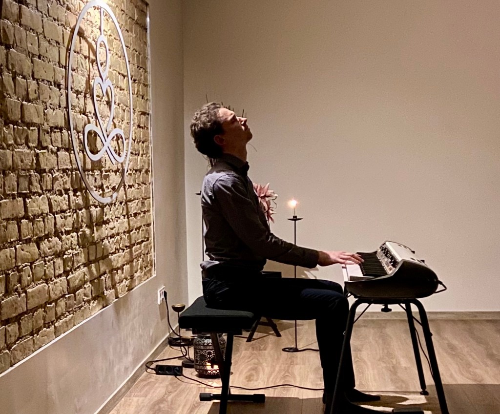 A man playing a keyboard during a meditation session in a minimalistic room with exposed brick walls, soft lighting, and a single lit candle.